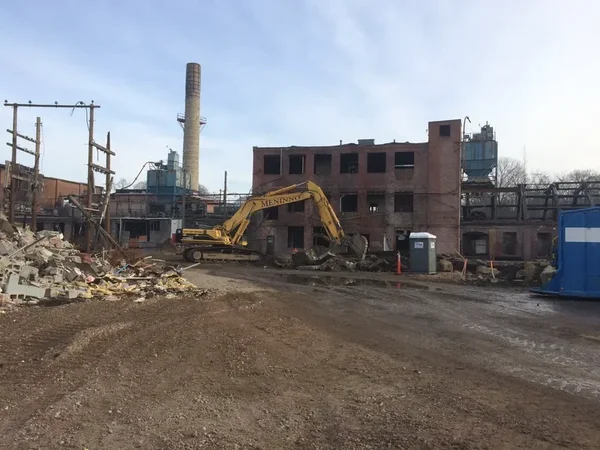 Demolition in progress at the Plymouth Rubber site — a yellow excavator working on a partially demolished brick building, smokestack in background.